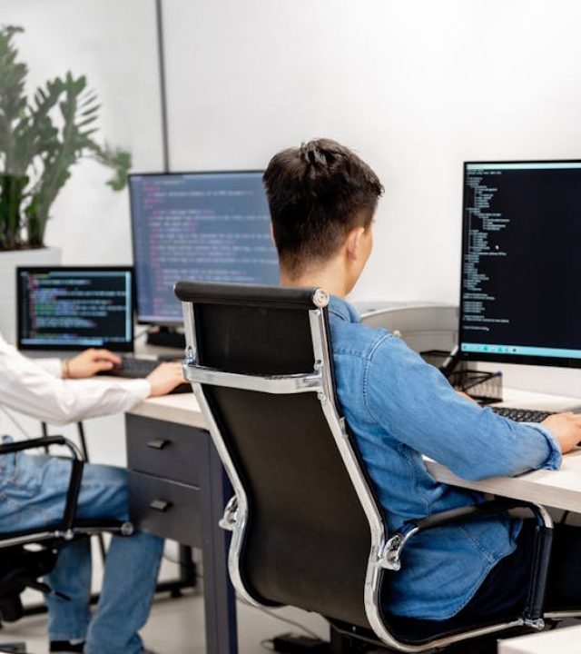 Two male developers at desks programming in a modern office workspace with large monitors.